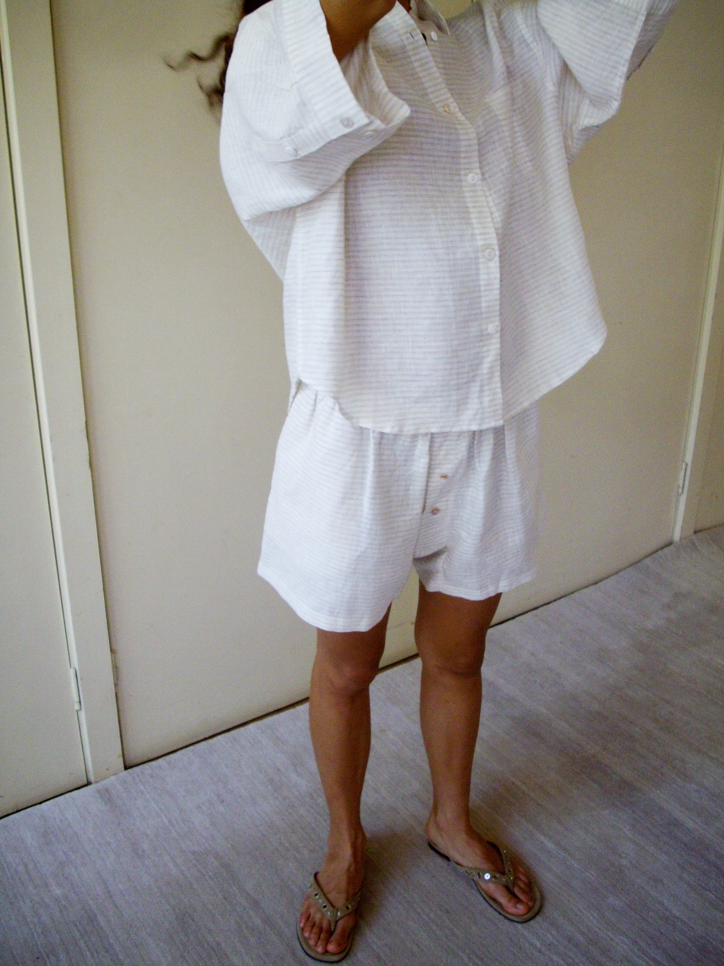 A person stands indoors on a light floor, wearing the 03 Set - Linen Story Stripe: a loose, long-sleeve organic linen shirt with matching boxer-style shorts. Their head is partially out of frame and they are also wearing sandals.