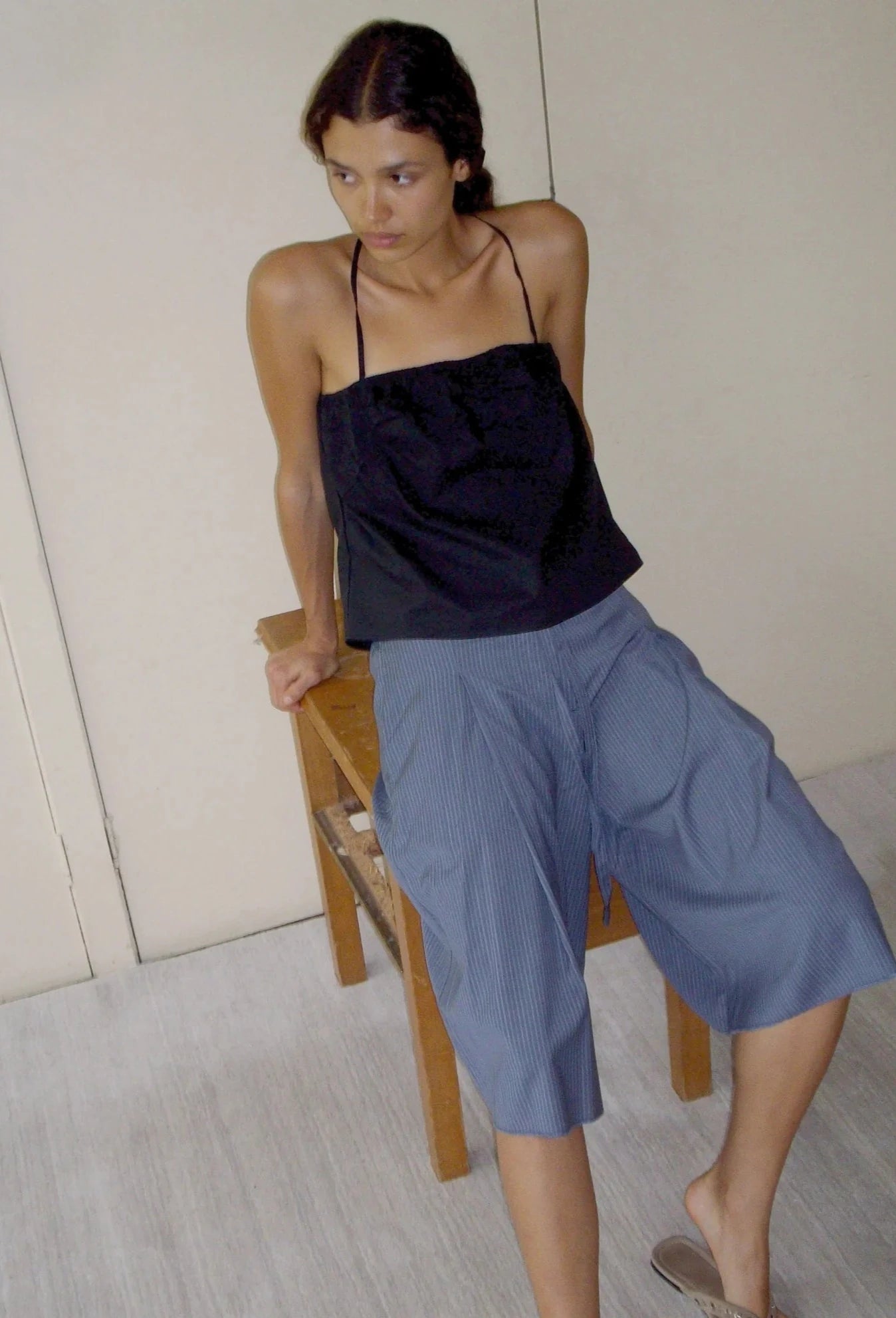 A person wears the Tube Top - Black by Deiji Studios and loose blue culottes, leaning against a wooden chair in a minimalist indoor space with neutral walls and light flooring.