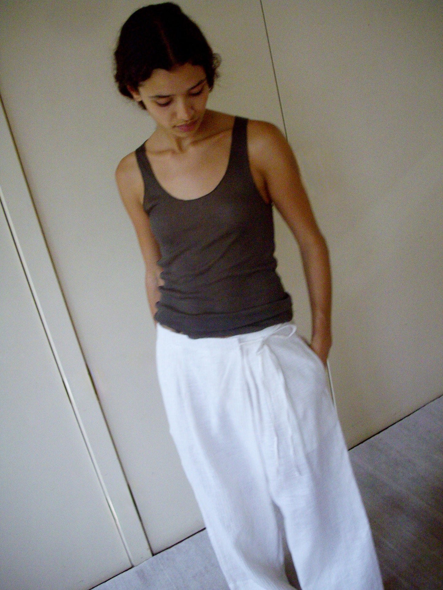 A person with short dark hair stands indoors against a beige background, looking down with hands in pockets, wearing the Knit Tank - Stone by Deiji Studios and loose white Ecovero viscose pants.