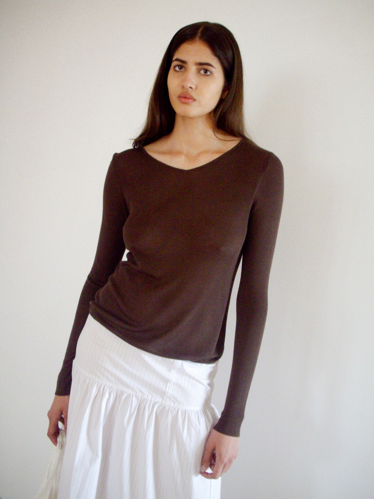 A woman with long dark hair wears the 0 Knitted Top - Dark Brown, a fitted, long-sleeved ecoviscose knit with sculptural seam details, styled with a white skirt as she stands against a plain light wall and calmly faces the camera.
