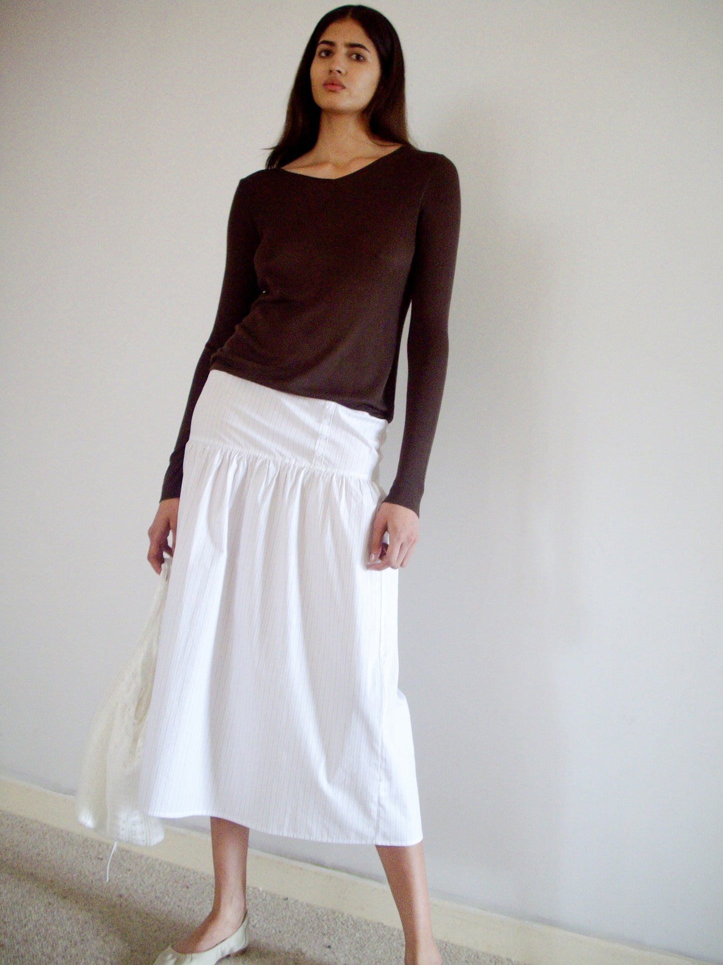 A woman with long dark hair stands indoors against a plain wall, wearing the 0 Knitted Top - Dark Brown, a long white skirt, and light-colored flats. She has a neutral expression.