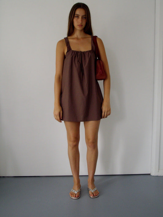 A woman with long brown hair and a neutral expression stands indoors against a plain white wall, wearing The Bow Dress - Cedar, silver sandals, and carrying a small red handbag.