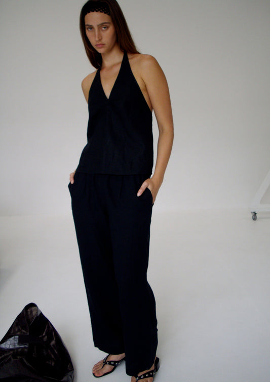A person stands in a minimalist room wearing a sleeveless black halter top and Linen Ease Trouser - Black, hands in pockets. They have long straight hair with a headband, wear black sandals, and a dark bag rests on the floor nearby.