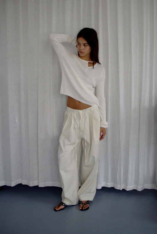 A woman stands before white curtains, wearing a long-sleeve white ribbed top and the Turn Pant - Off White by Deiji Studios, featuring a relaxed tailored fit and adjustable drawcord. She gazes to the side with one arm resting on her head.