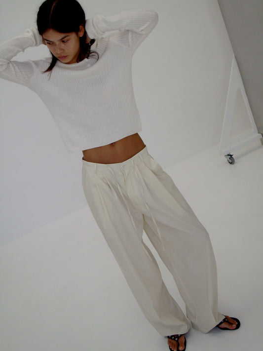 A person stands indoors in a white long-sleeve shirt, Turn Pant - Off White by Deiji Studios featuring an adjustable drawcord, and black sandals, arms raised behind their head while looking down against a minimal white background.