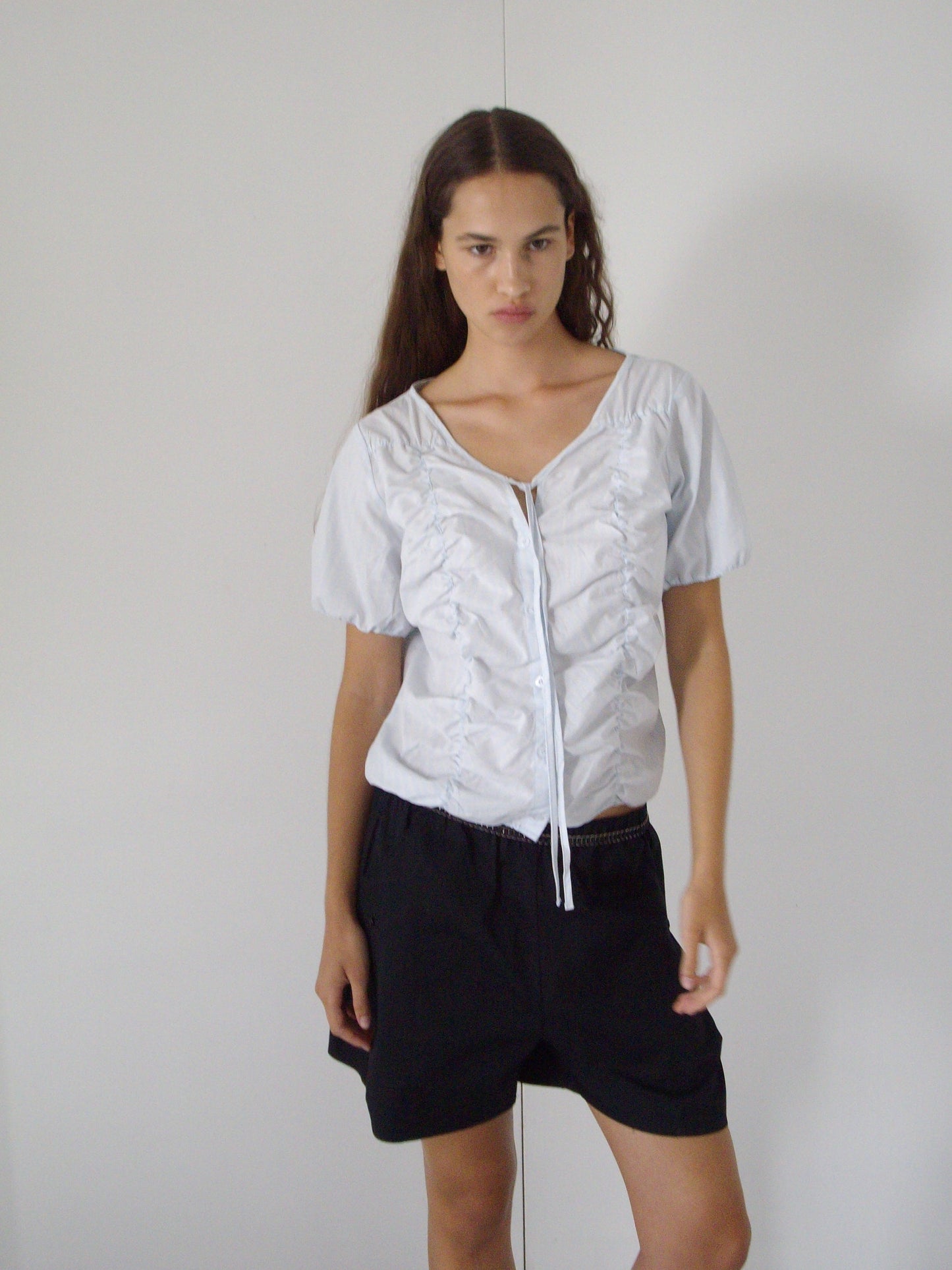 A person with long brown hair wearing the Crinkle Top - Powder Blue Stripe and black shorts stands in front of a plain white wall, looking slightly to the left.
