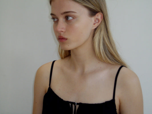 A young woman with long blonde hair, wearing the Lettuce Mini Dress - Black by Deiji Studios, looks to her left against a plain light background.