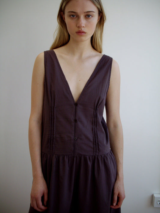 A woman with long blonde hair wears the Pintuck Dress - Raisin from Deiji Studios, a sleeveless, deep V-neck, dark mid-length dress, and stands against a plain white wall looking directly at the camera with a neutral expression.