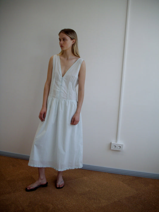 A woman stands indoors against a plain white wall, wearing the Pintuck Dress - Mint by Deiji Studios with a deep V-neck and flat sandals. She gazes slightly to the side with a neutral expression.