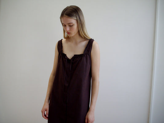 A woman with long, straight hair stands against a plain white wall, wearing the sleeveless Button Sweep Dress - Umber by Deiji Studios, featuring subtle ruffle details at the neckline. She looks down with a calm expression.
