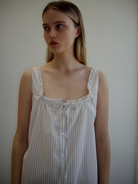 A young woman with long, straight, light brown hair stands against a plain background, wearing the Button Sweep Dress - Gull Stripe. The relaxed style features ruffled edges and front buttons as she gazes to the side.
