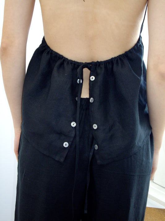 A person wears the Linen Button Halter - Black by Deiji Studios, a backless linen top with adjustable drawstring and button details down the back, exposing their upper and mid-back; their arms rest relaxed at their sides.