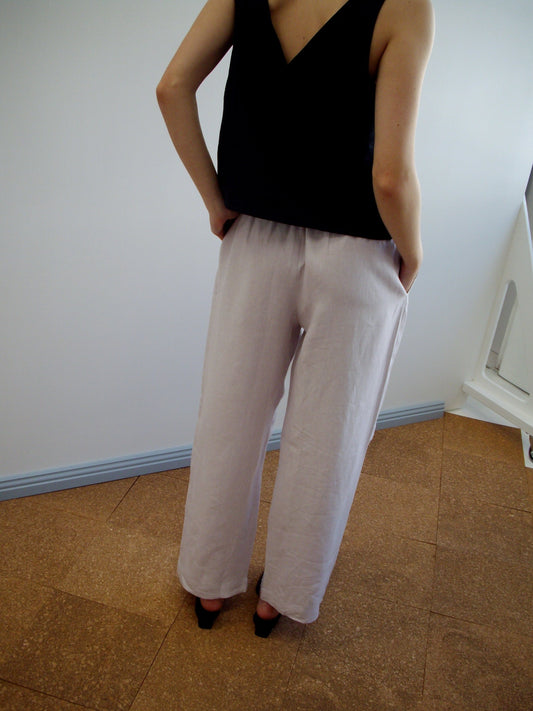 A person stands indoors, back turned, wearing a sleeveless black top and Linen Ease Trouser - Lilac with an elastic waist and straight leg fit. Hands in pockets, black heeled shoes. White and blue walls with brown flooring in the background.