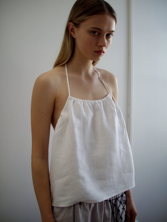 A person with long, straight blonde hair wears the O Ring Halter - White from Deiji Studios with matching pants, standing indoors against a plain white wall and looking slightly to the side with a neutral expression.