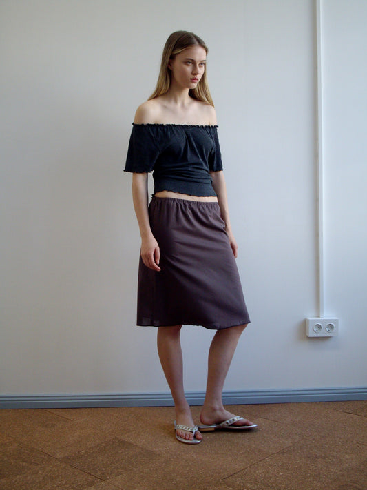 A woman with long, straight hair and a neutral expression stands indoors against a plain wall, wearing an off-the-shoulder black top, sandals, and the Simple Skirt - Raisin.