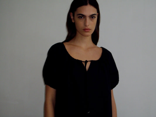 A model with long dark hair wears the Smock Dress - Black by Deiji Studios, featuring puff sleeves and a keyhole neckline with a small tie detail, photographed against a light grey background.
