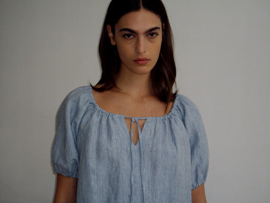 A woman with long brown hair stands against a plain light background, wearing a light blue, short-sleeved Smock Dress with a front tie, looking directly at the camera with a neutral expression.