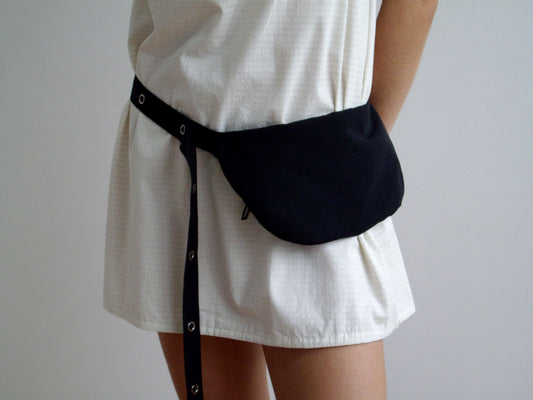 Wearing a short, white textured dress, the person styles the Snap Belt Bag - Black from Deiji Studios at their waist, posed against a plain light background to highlight this belt bag as an essential everyday accessory.
