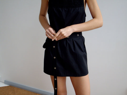 Person stands indoors in a sleeveless black dress, fastening the Snap Belt Bag - Black around their waist. The everyday accessory features silver eyelets. Background is plain and light-colored.