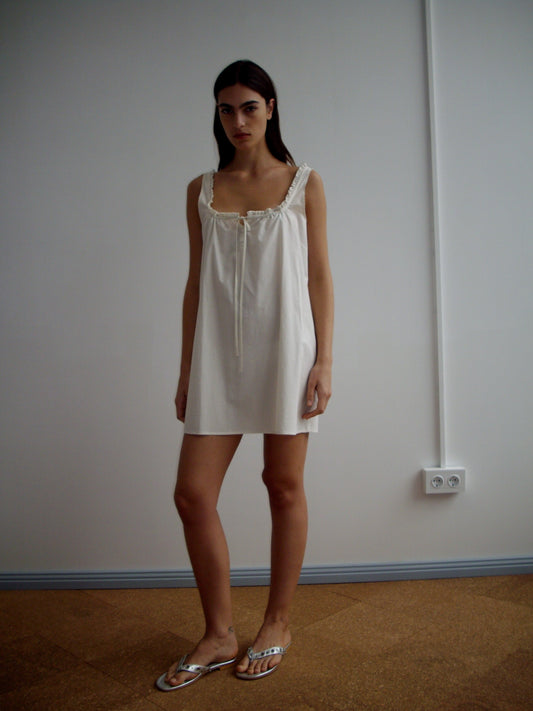 A woman with long dark hair stands indoors against a plain white wall, wearing the Button Frill Mini Dress - Stone Stripe. She also has on metallic flat sandals, and an electrical outlet is visible on the wall behind her.
