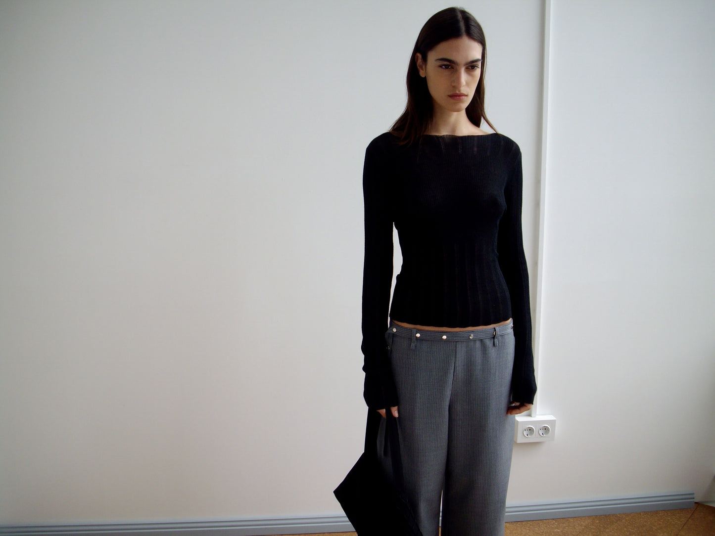 A woman with straight dark hair wears a fitted black long-sleeve top and Wool Belt Pant in Grey Blue Stripe, standing indoors against a plain white wall while holding a black bag at her side.
