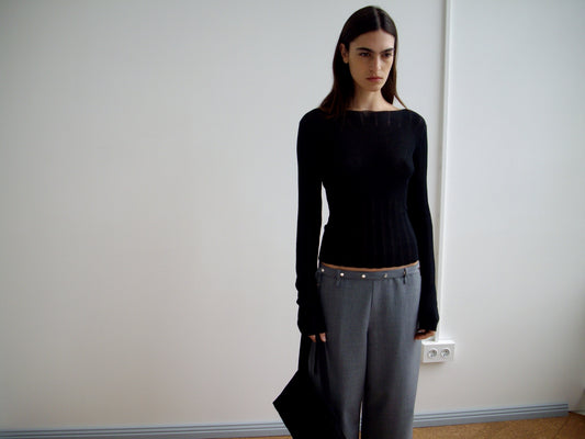 A person with long dark hair stands indoors against a white wall, wearing a fitted black sweater and Deiji Studios' Wool Belt Pant in Grey Blue Stripe with a visible waistband. They hold a black bag and have a neutral expression.
