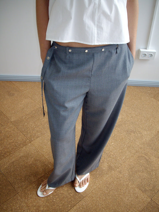 A person stands on a brown tile floor against a plain wall, wearing Deiji Studios' Wool Belt Pant in Grey Blue Stripe with side button details, paired with a white sleeveless top and white sandals.