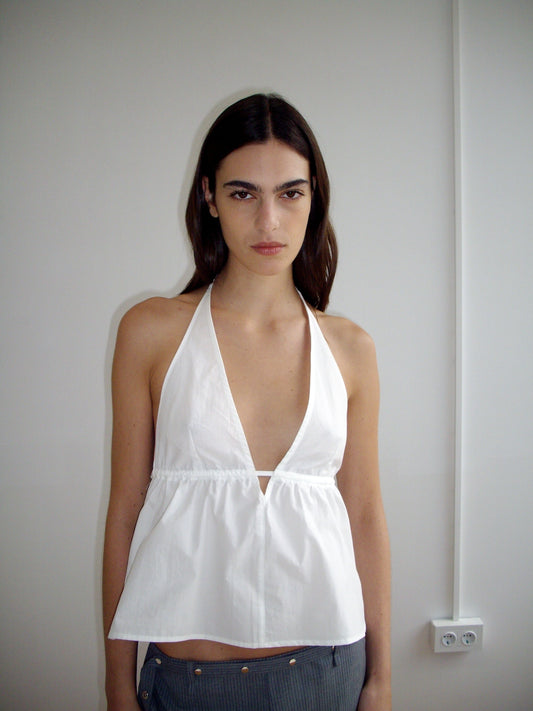 A person with long dark hair wears the Button Halter - White by Deiji Studios, standing against a light-colored wall with a power outlet and visible wire in the background.
