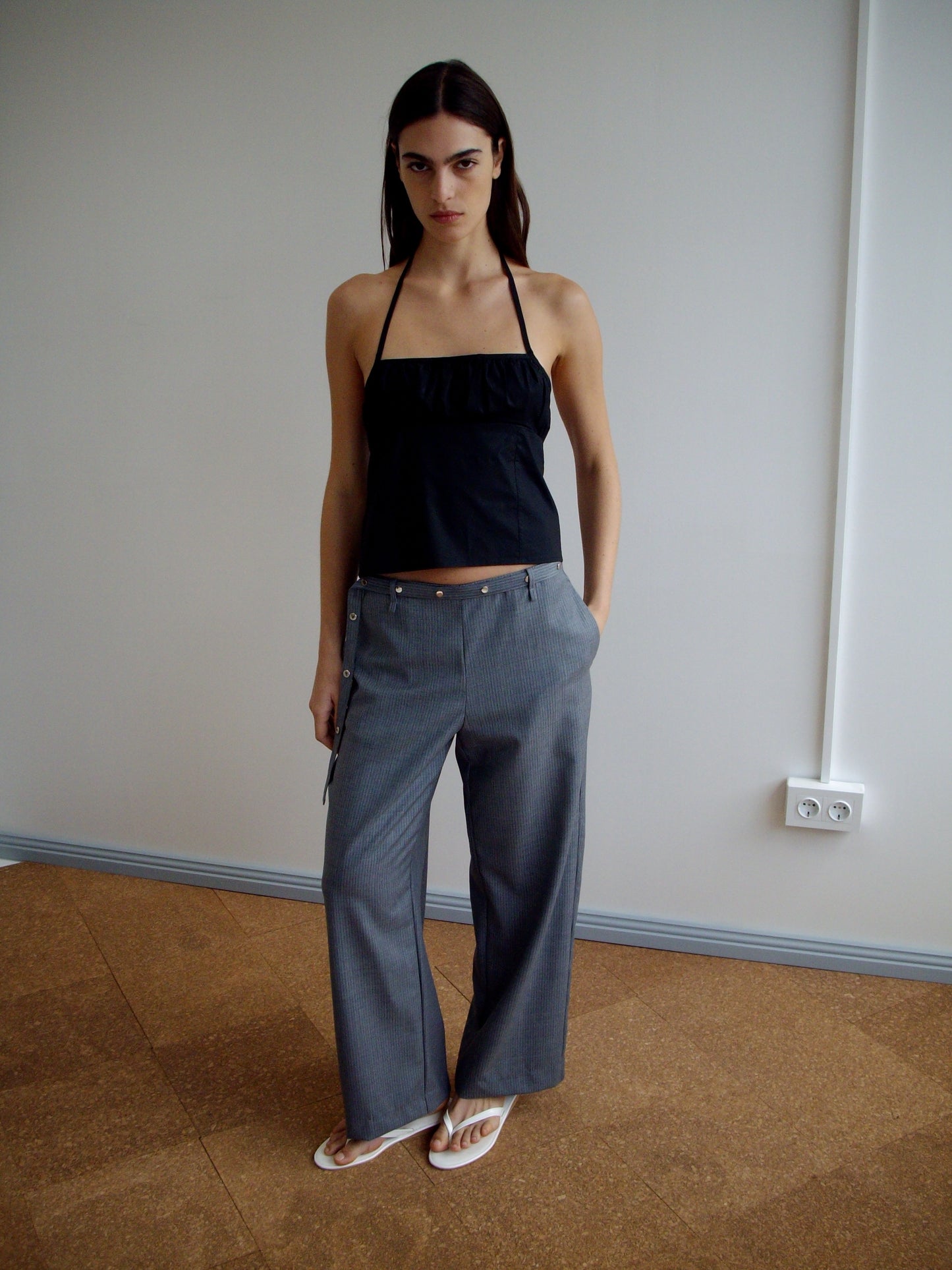 A woman stands indoors against a plain wall, wearing a black halter top and Wool Belt Pant - Grey Blue Stripe with white sandals. She has long dark hair, a serious expression, and her hands in the pockets of her pants.