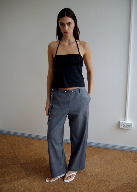 A person stands indoors wearing the Deiji Studios Strap Top - Black, loose gray zippered trousers, and white flip-flops against a simple backdrop of a white wall and brown floor.