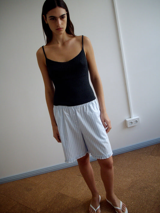 A person with long dark hair stands indoors against a white wall and brown floor, wearing a black tank top, Flounce Short - Mineral Stripe by Deiji Studios, and white flip-flops.
