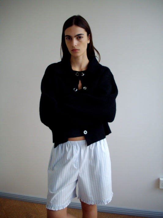 A person with long dark hair stands against a plain wall, arms crossed and serious, wearing a black buttoned sweater and the Flounce Short - Mineral Stripe from Deiji Studios.