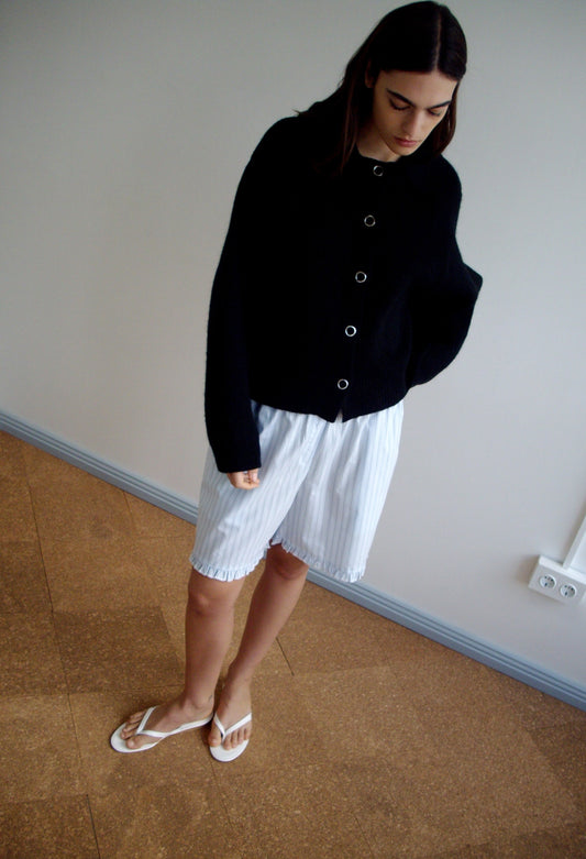 A woman stands indoors on a brown floor, wearing the Stud Cardi Knit - Black, loose white striped shorts, and white sandals. She looks down with her hands in her pockets against a plain light wall.
