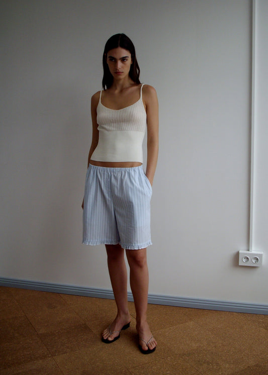 A woman stands indoors against a plain wall, wearing a Ribbed Tank - Off White from Deiji Studios, light blue striped shorts, and brown sandals. She has long straight hair, a neutral expression, and her hands in her pockets.