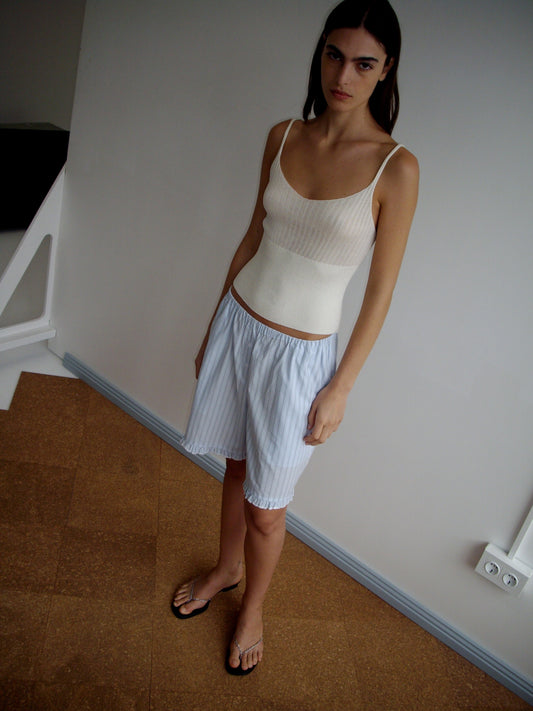 A woman with long dark hair stands indoors on a brown and white floor, wearing a Deiji Studios Ribbed Tank in Off White, loose light blue striped shorts, and flip-flops. She looks at the camera with a neutral expression.