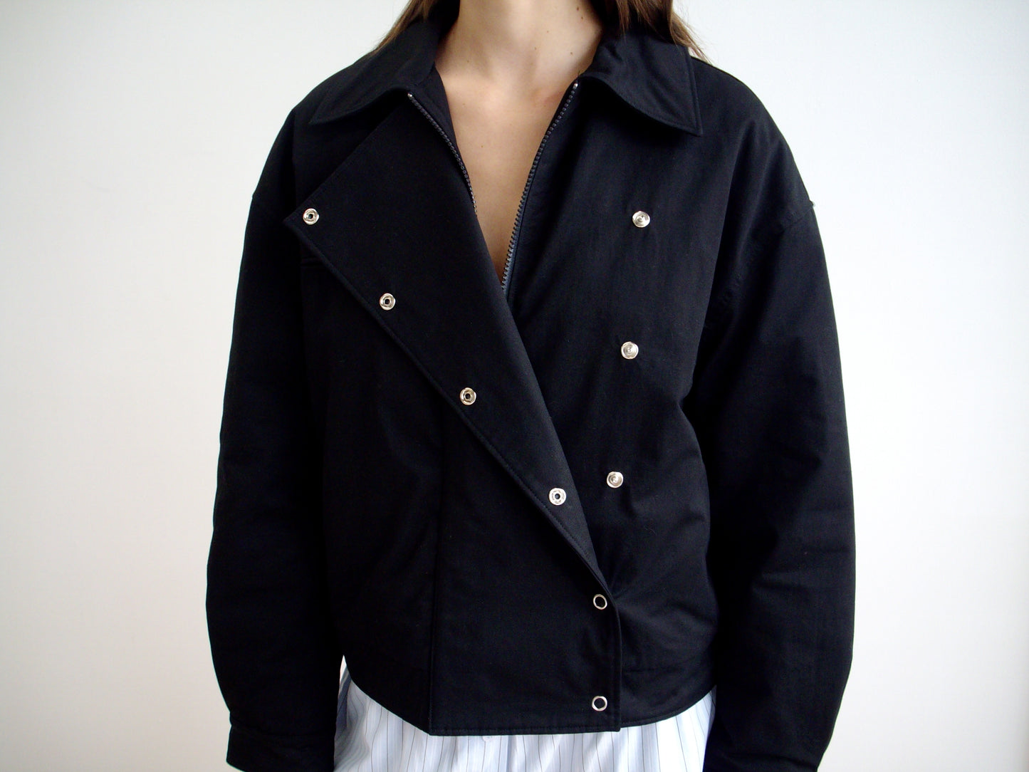 A person models the Lapel Jacket - Black with asymmetric lapels and shiny silver buttons, partially unzipped over a white outfit against a plain light background, highlighting its V neckline.