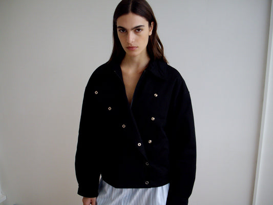 A person with long dark hair stands indoors against a plain light wall, wearing the Deiji Studios Lapel Jacket - Black and light-colored pants, looking directly at the camera with a serious expression.