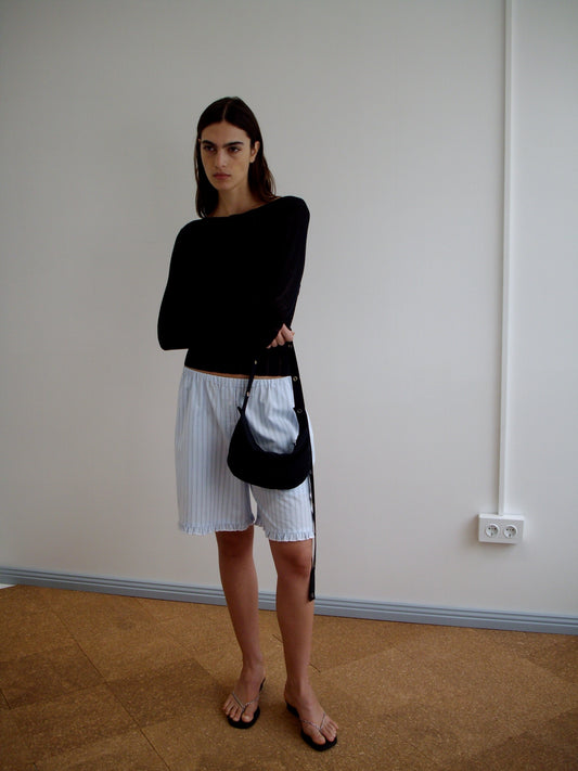 A woman stands indoors against a plain white wall, wearing a black long-sleeve top, loose white pinstriped shorts, flip-flops, and carrying the Snap Belt Bag - Black—an effortless everyday accessory. She has long dark hair and a neutral expression.