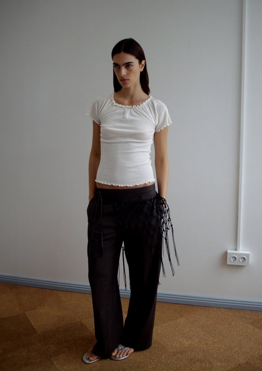 A model wears the Flounce Tee - Cream by Deiji Studios with a ruffled neckline and hem, styled with dark wide-leg trousers featuring black ribbon side details and silver sandals.
