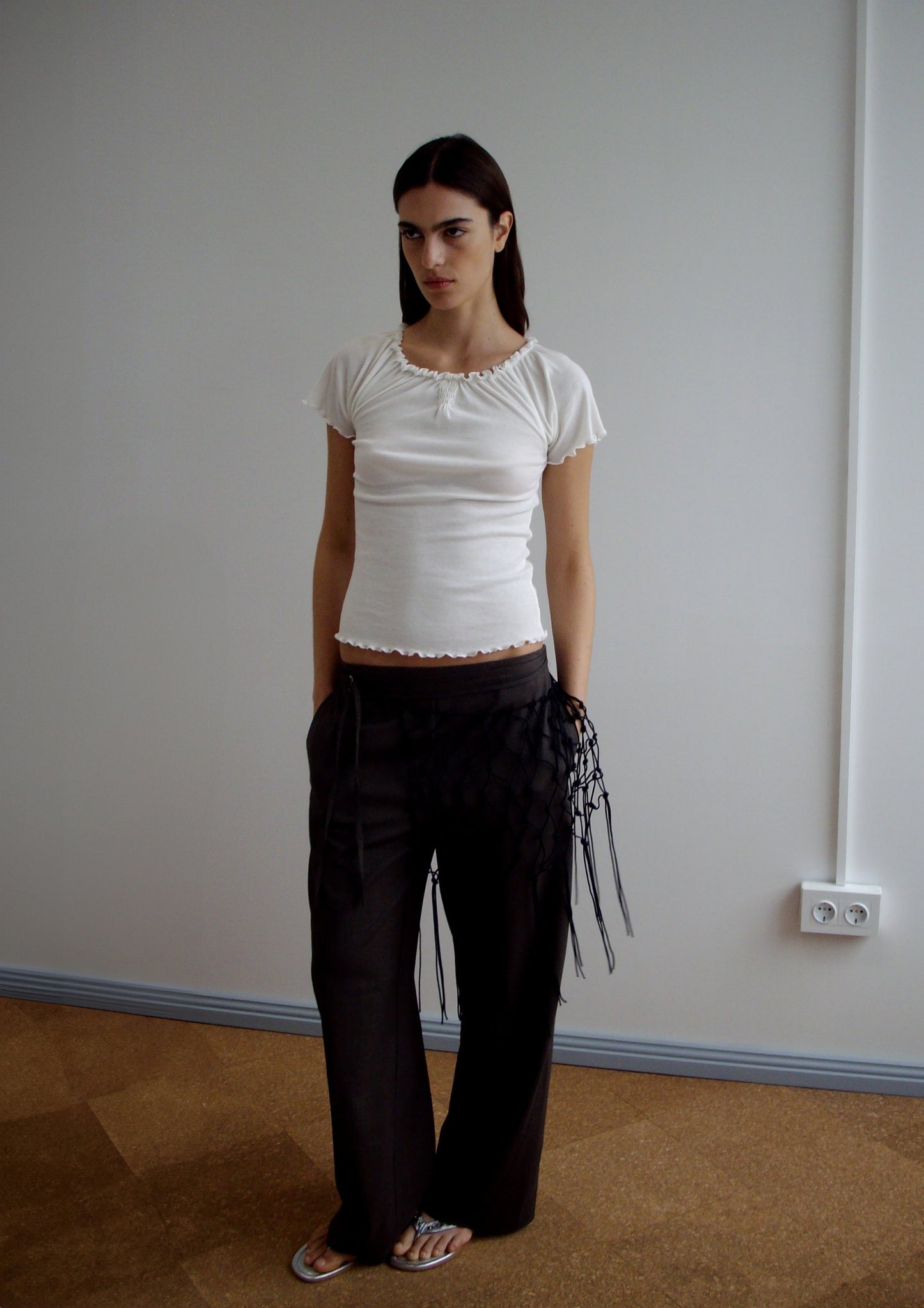 A woman with long dark hair and a neutral expression stands indoors against a plain wall, wearing the Flounce Tank - Cream from Deiji Studios, loose dark pants with netted fringe detail, and white flip-flops.
