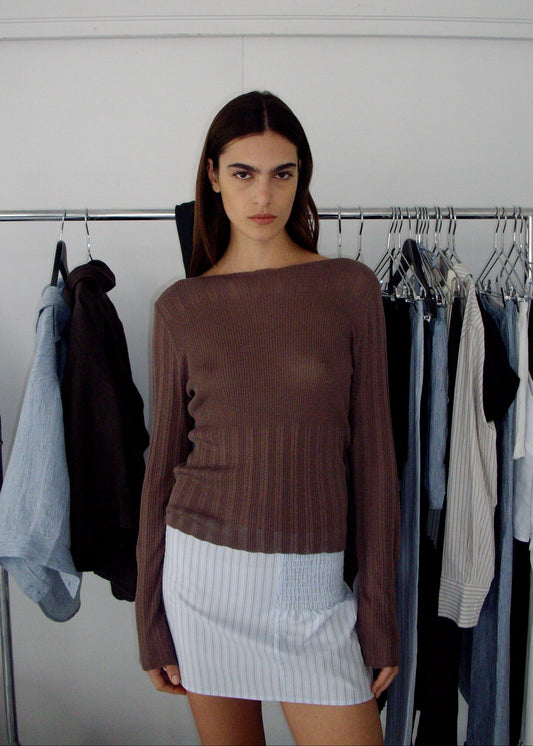 A woman with long dark hair stands before a clothing rack, wearing a brown ribbed top and the Shirring Skirt - Mineral Stripe, featuring light blue vertical stripes, while various garments hang behind her.