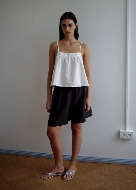 A woman stands indoors against a plain wall, wearing a white Yoke Top from Deiji Studios, loose dark shorts, and sandals. She has straight dark hair and a neutral expression. The floor is tan and there's an outlet on the wall.