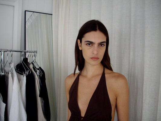 A person with long dark hair and a serious expression wears the Button Halter - Umber, a deep v-neck brown halter top with an adjustable backline, standing indoors by a clothing rack and a full-length mirror framed by white curtains.