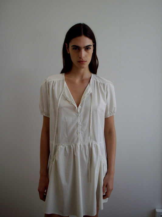 A woman with long dark hair stands against a white wall, wearing the Pocket Mini Dress—a light short-sleeved, button-front dress with a loose, relaxed fit. She gazes at the camera with a neutral expression.