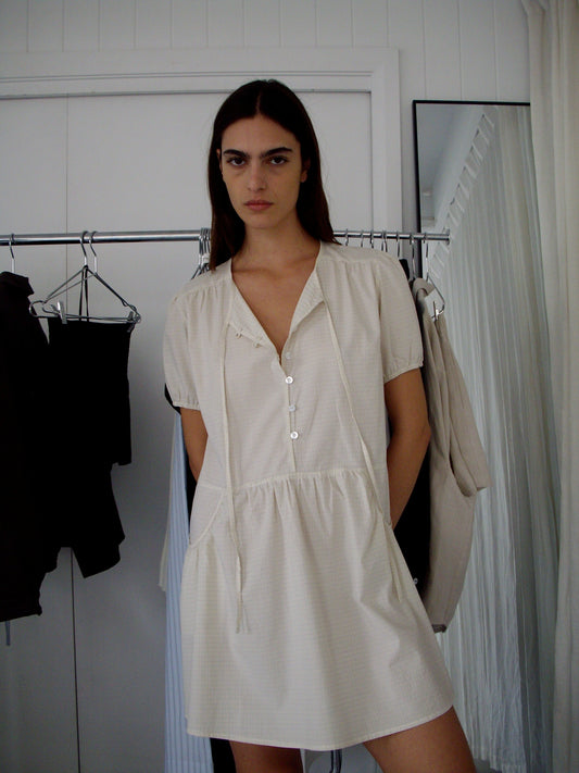 A woman with long dark hair stands indoors in front of a clothing rack and white paneled wall, wearing the Pocket Mini Dress - Bone Check.