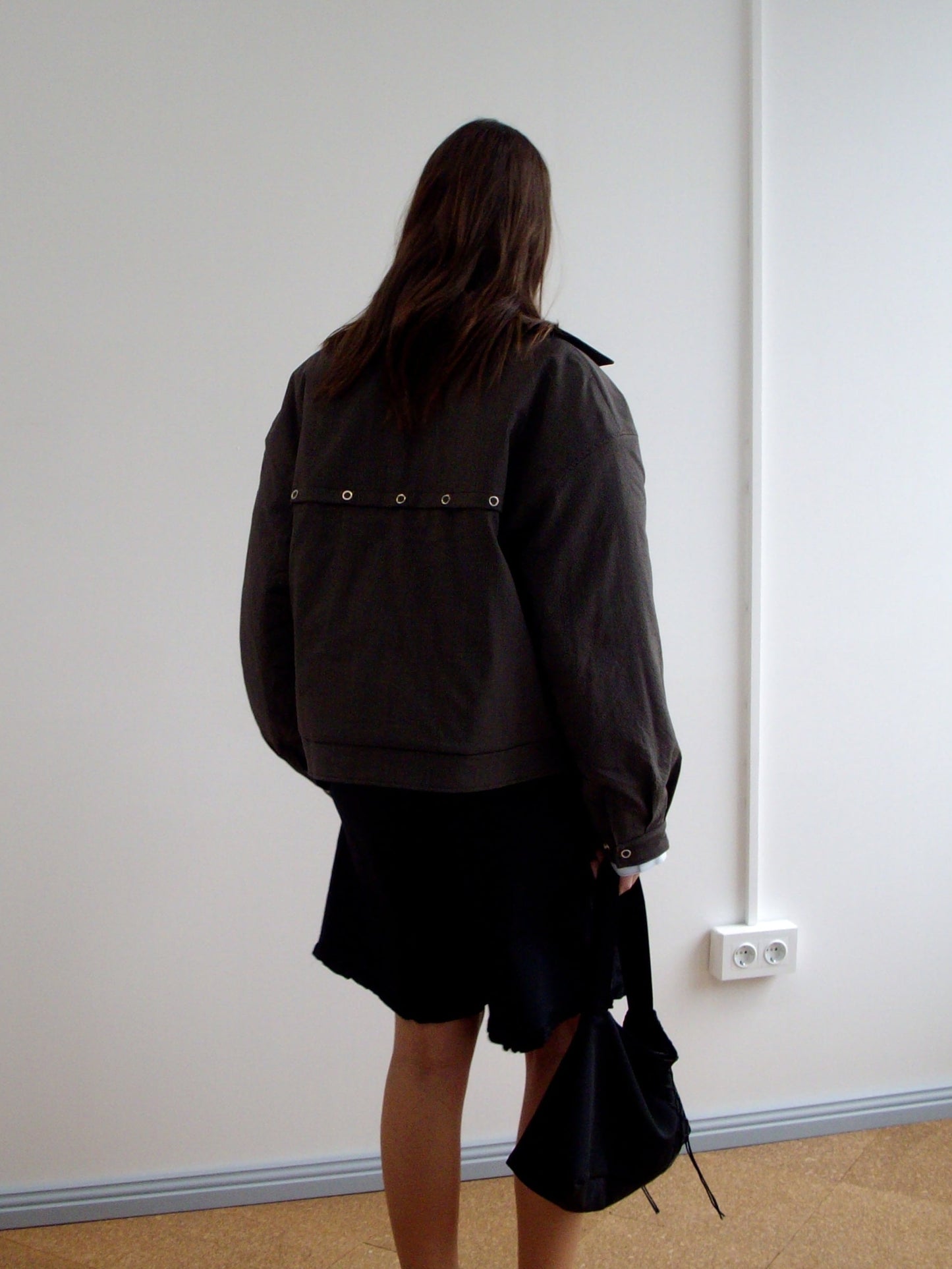 A person with long brown hair stands facing a white wall, wearing the Lapel Jacket - Brown Stripe from Deiji Studios and a black skirt with an uneven hem, holding a black bag in their right hand.