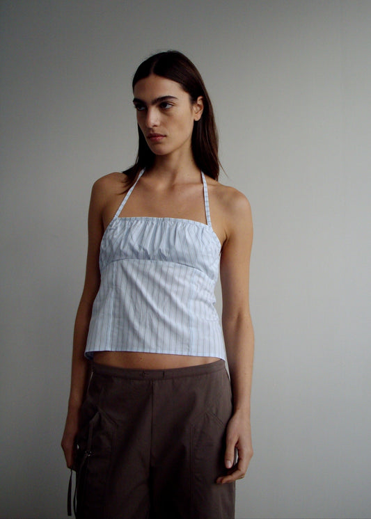 A woman stands against a plain background wearing the Strap Top - Mineral Stripe by Deiji Studios and brown pants, looking slightly to the side with a neutral expression.