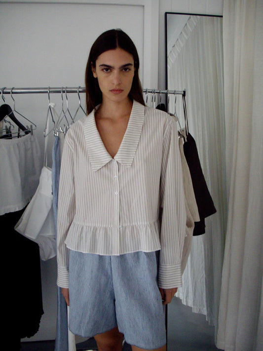 A model wears a white and blue stripe peplum blouse with an oversized collar and the Press Stud Short - Blue Stripe, posing in front of a clothing rack in a minimalist studio setting.