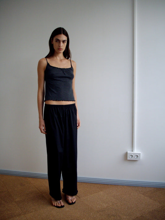 A model wears the Wool Blend Flounce Tank - Grey by Deiji Studios with black wide-leg drawstring trousers featuring ruffled hems and strappy sandals, standing against a light grey wall.
