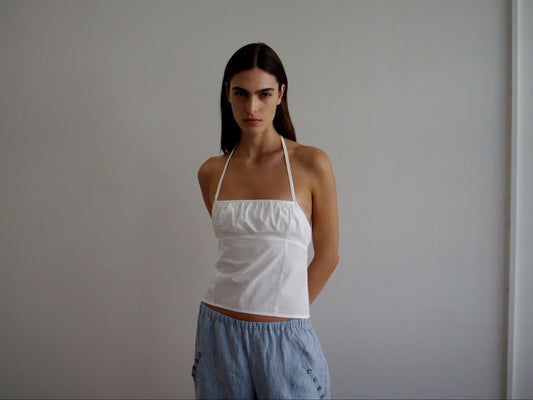 Model wearing the Strap Top - White by Deiji Studios, featuring a square neckline and gathered bust, paired with light blue striped wide-leg trousers with button accents, set against a grey background.